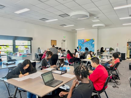 Students sitting at tables in a classroom working on laptops and writing on papers