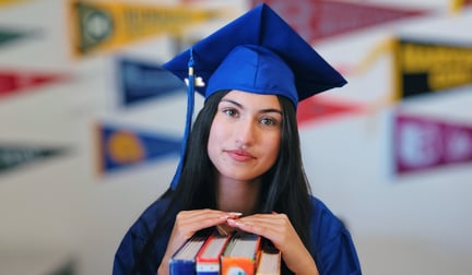 A student in a blue graduation cap and gown is sitting at a desk with textbooks and posing in front of a wall of college pennants