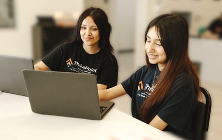 Two students sitting at a table, smiling and working on laptops together