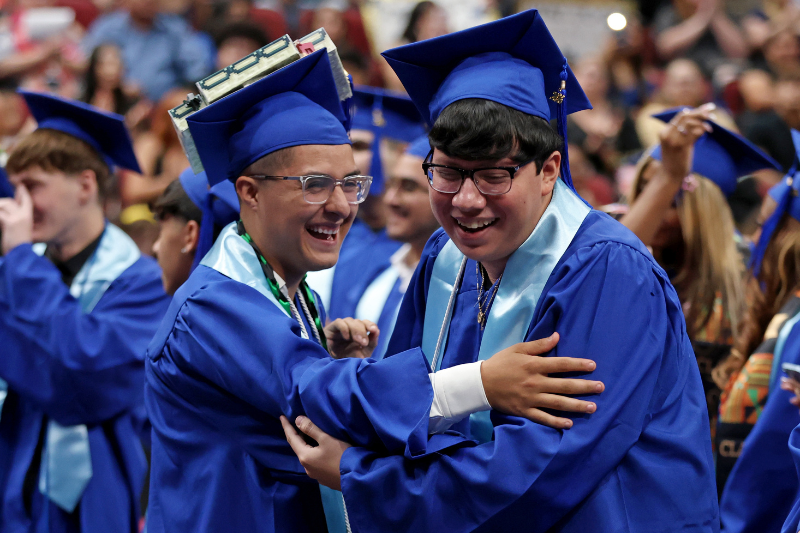 two students wearing blue graduation cap and gowns celebrating and hugging